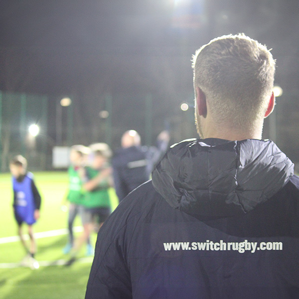 Switch Rugby coach leading youth rugby training session on floodlit field, developing skills and teamwork