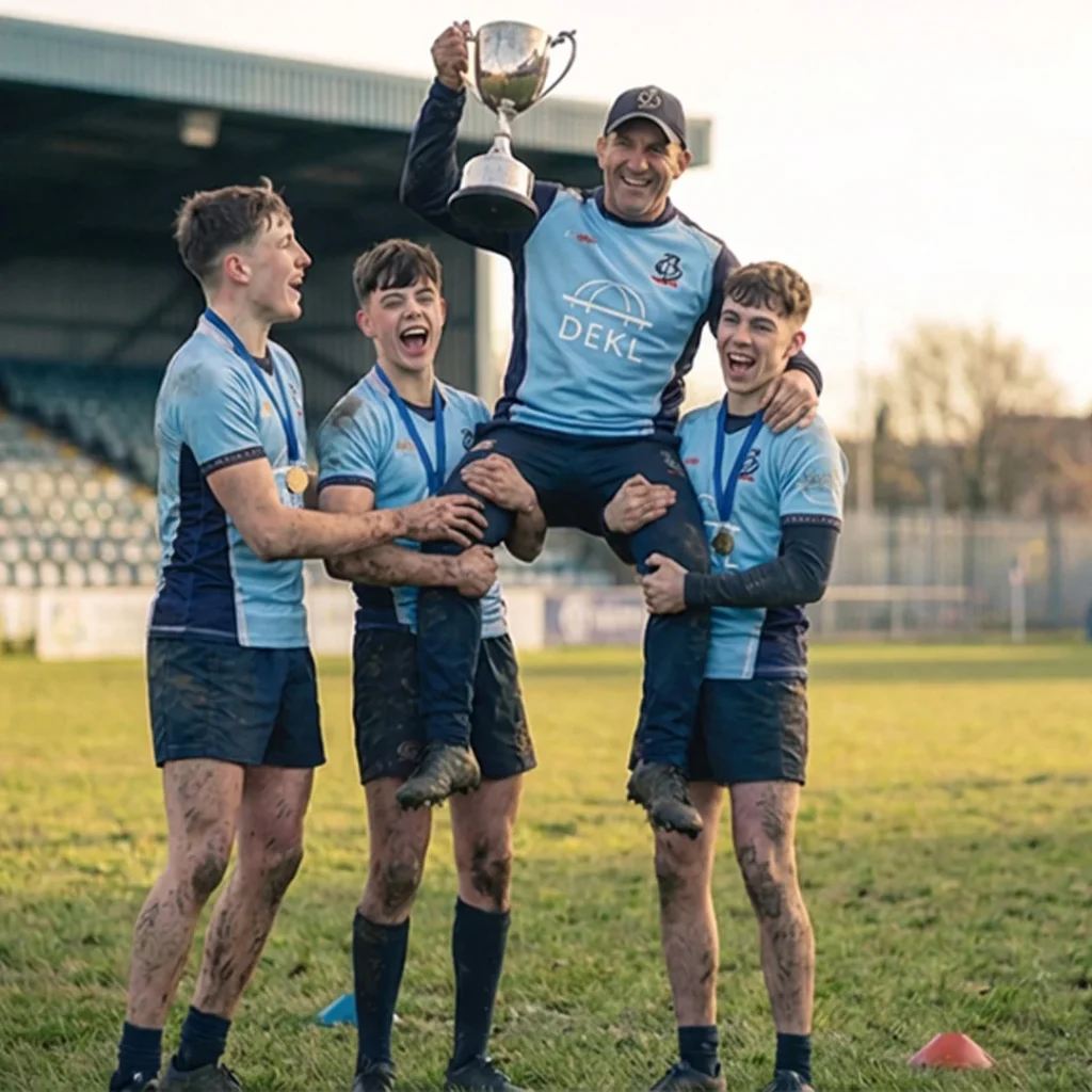 City of Bristol Rugby U16s players triumphantly lifting coach on shoulders while holding trophy – joyful victory celebration with medals and muddy kit