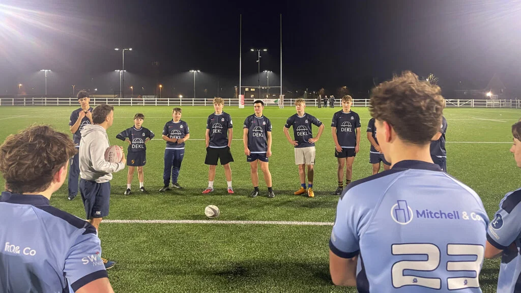 Night training session for Switch Rugby's City of Bristol Under 16s youth rugby programme at floodlit rugby pitch, teenage players in navy blue DEKL and Switch Rugby sponsored kits listening to coach, Bristol UK