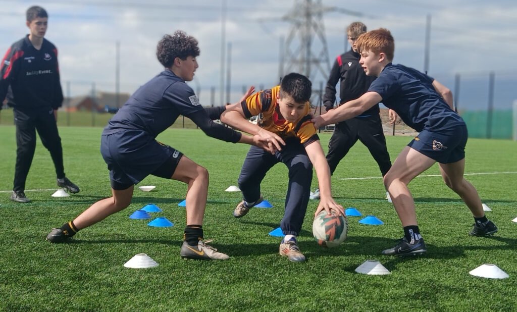 Three young rugby players contest the ball during a Switch Rugby Academy training drill on a 3G pitch